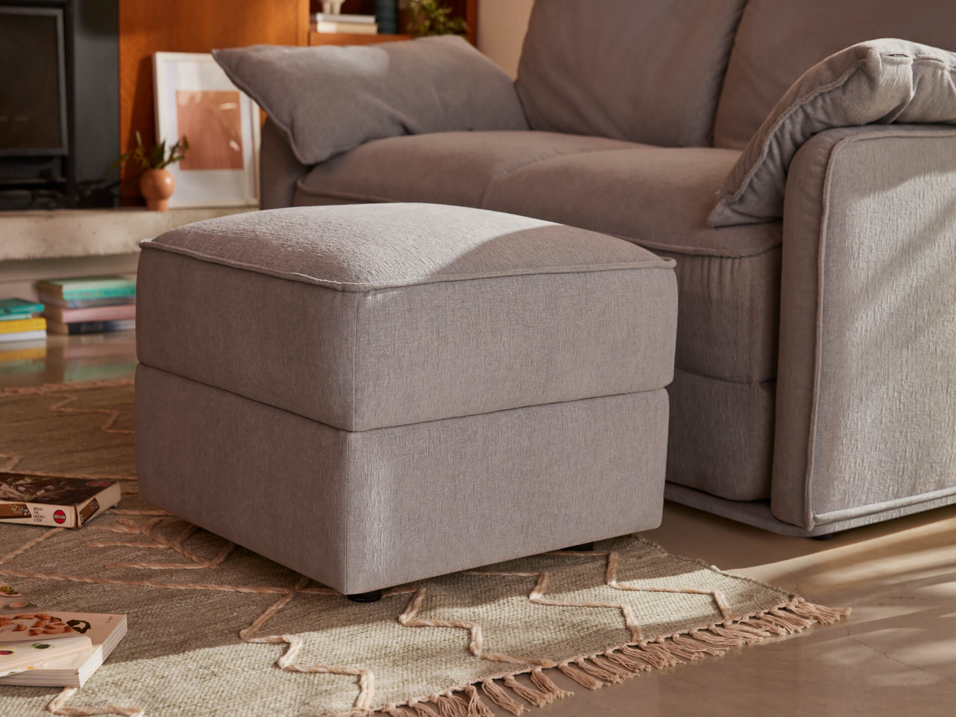 Grey ottoman beside matching sofa on a patterned rug, with books nearby, in a cosy living room.