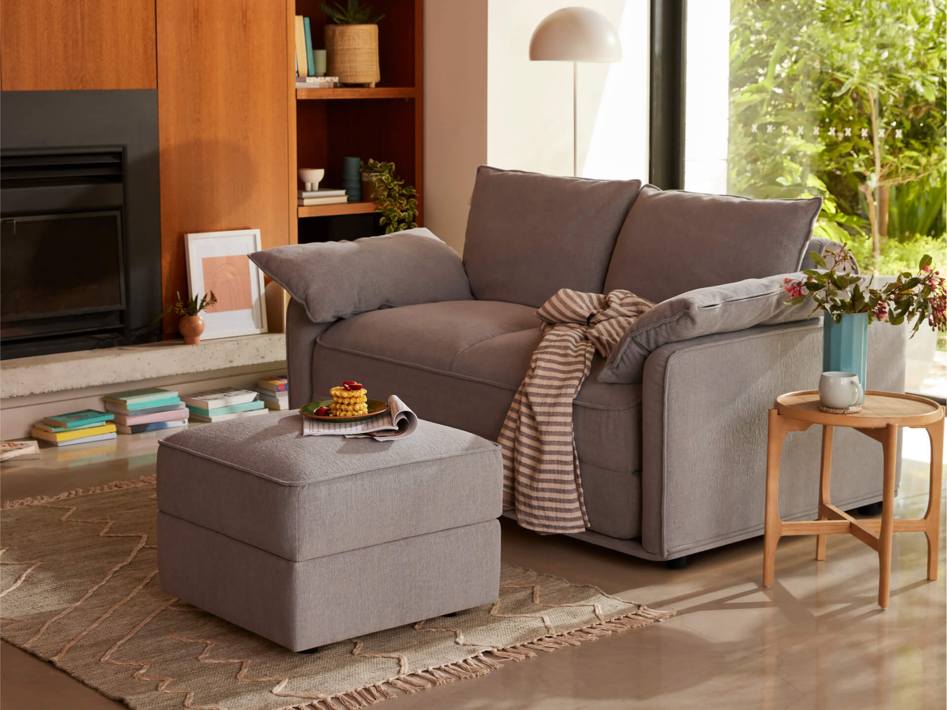 Grey couch with matching ottoman in a cosy living room, featuring a wooden side table, potted plants, and books on the floor.