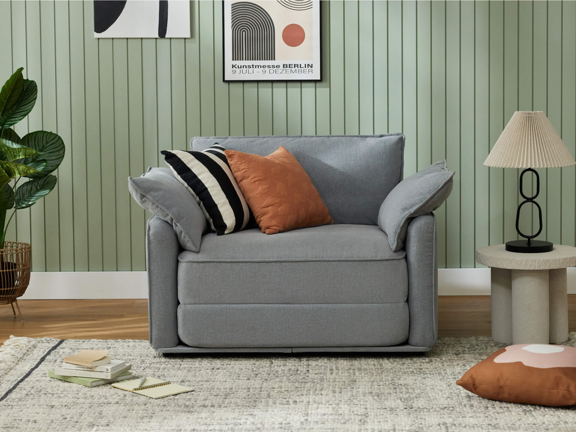 Grey armchair with orange and striped cushions in a living room. Green panelled wall, abstract art, round side table with lamp, and plant decor.