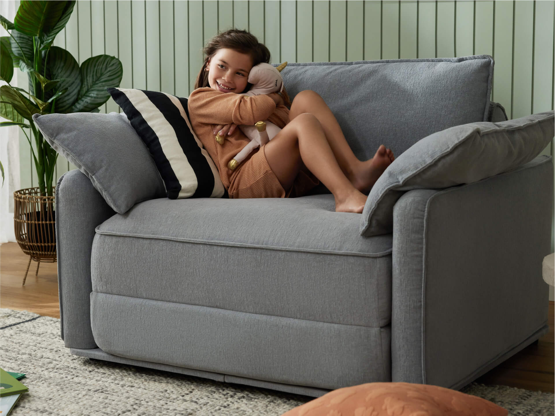 A young girl relaxes on a light grey armchair with plush cushions, hugging a stuffed toy. A striped cushion and plants are in the background.