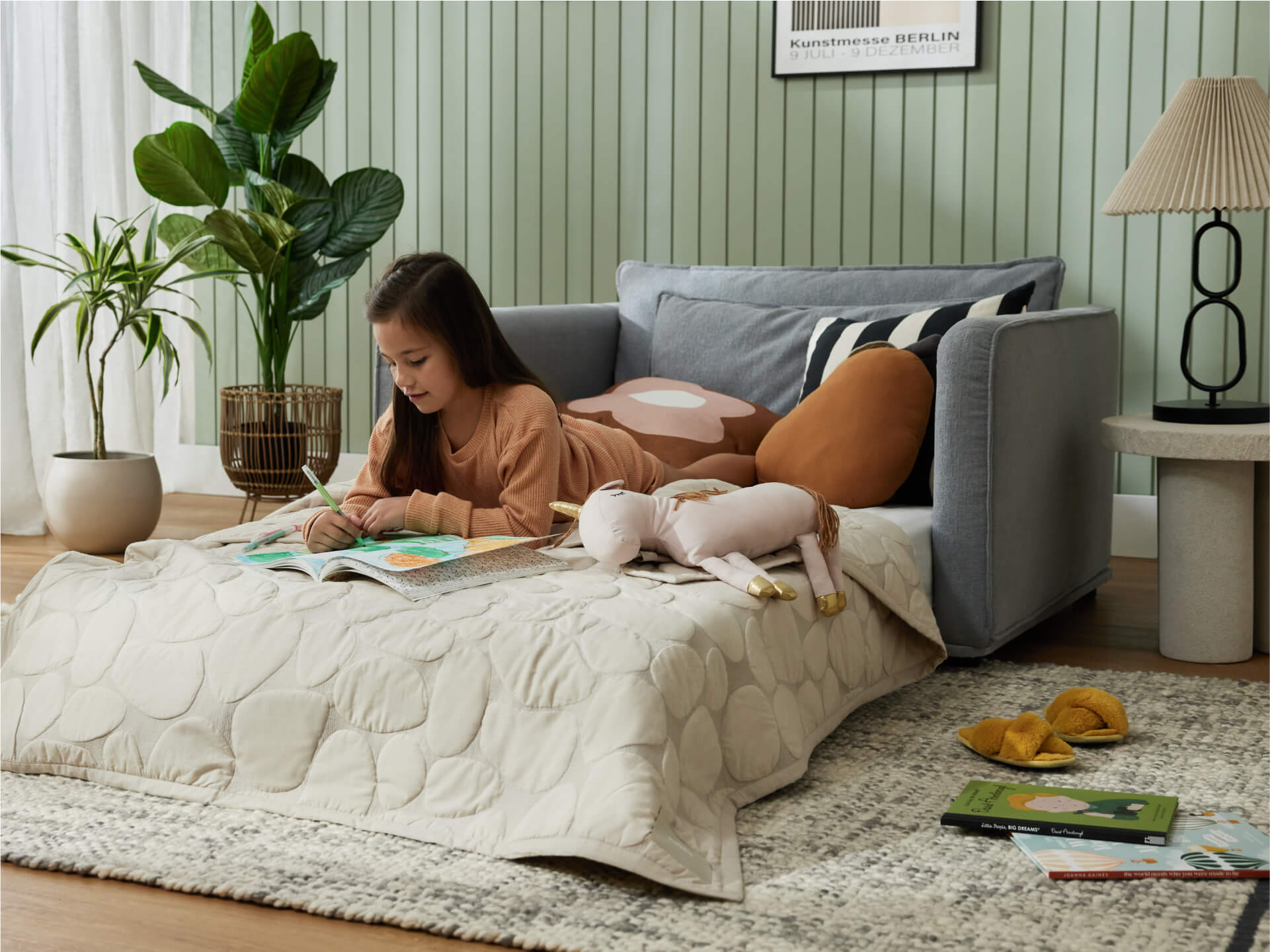 Girl reading on a light grey sofa bed with cushions, beige quilt, plush toy, and green plant. Room has a striped wall, lamp, and books on rug.