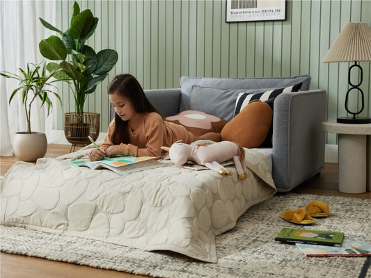 Girl reading on a beige quilted mattress with colourful pillows. Nearby is a grey sofa, a side table with a lamp, and potted plants.
