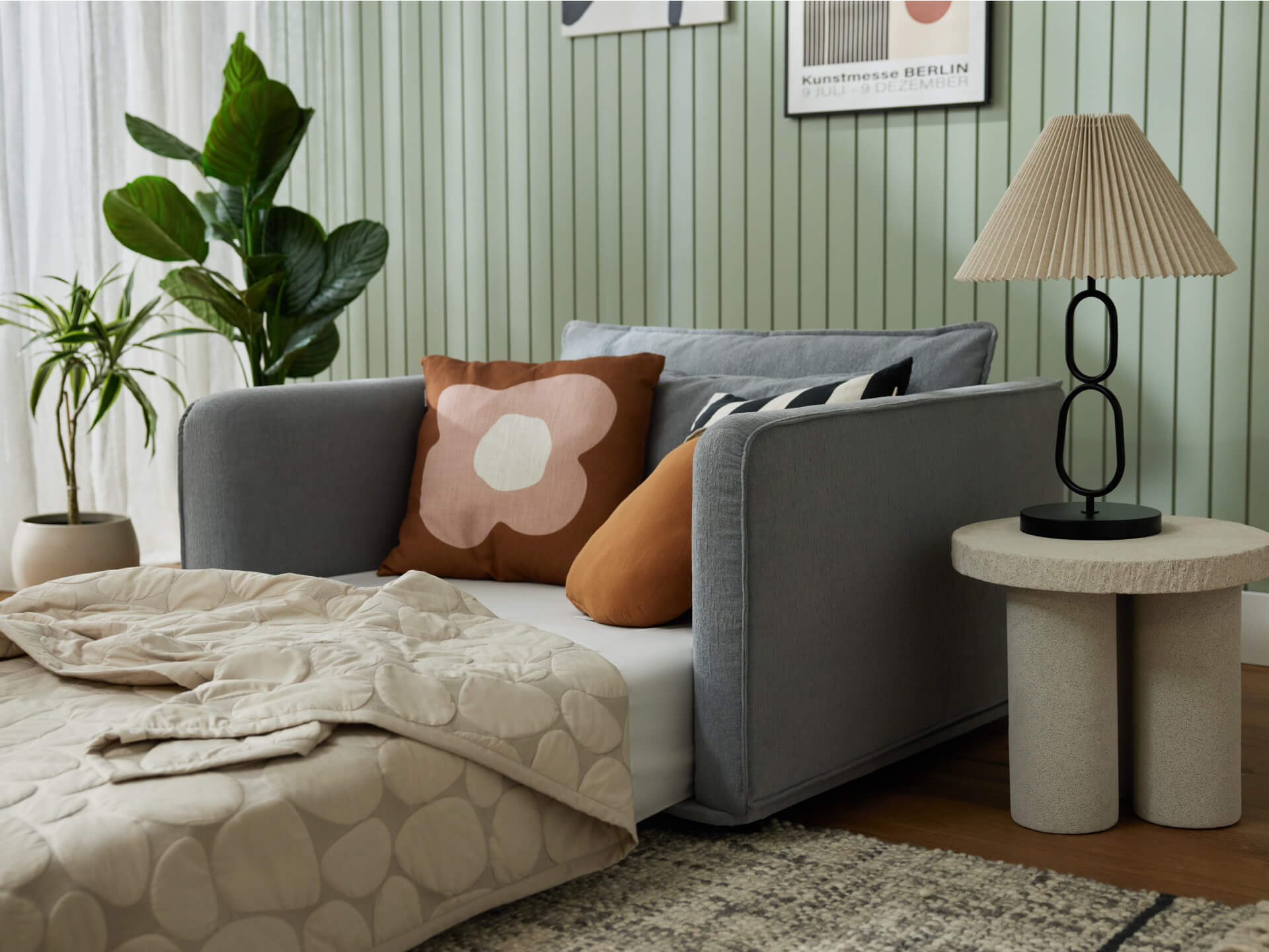 Cosy living room with grey sofa bed, patterned cushions, and beige quilt. Potted plants and a round table with a lamp complete the setting.