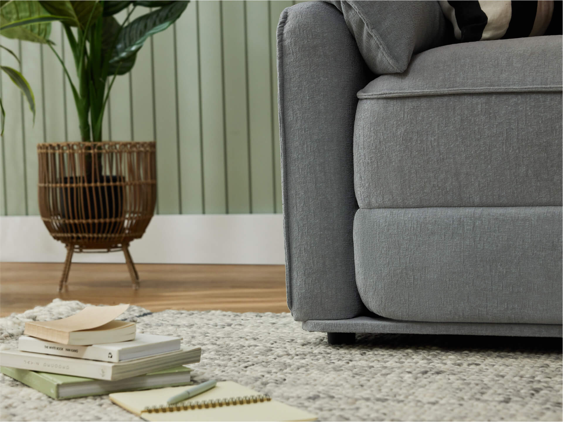 Close-up of a light grey sofa with a textured cushion on a grey rug. A pile of books and a notebook sit nearby, with a potted plant in the background.