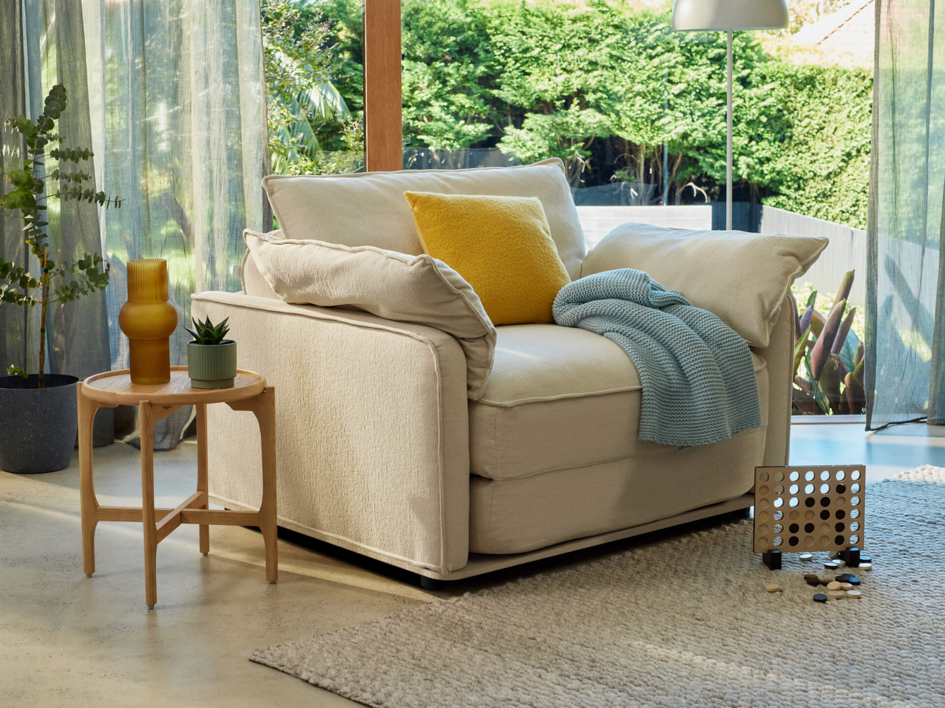 Beige armchair with yellow and blue cushions, near wood table with vase. Set in a bright room with large windows overlooking greenery.