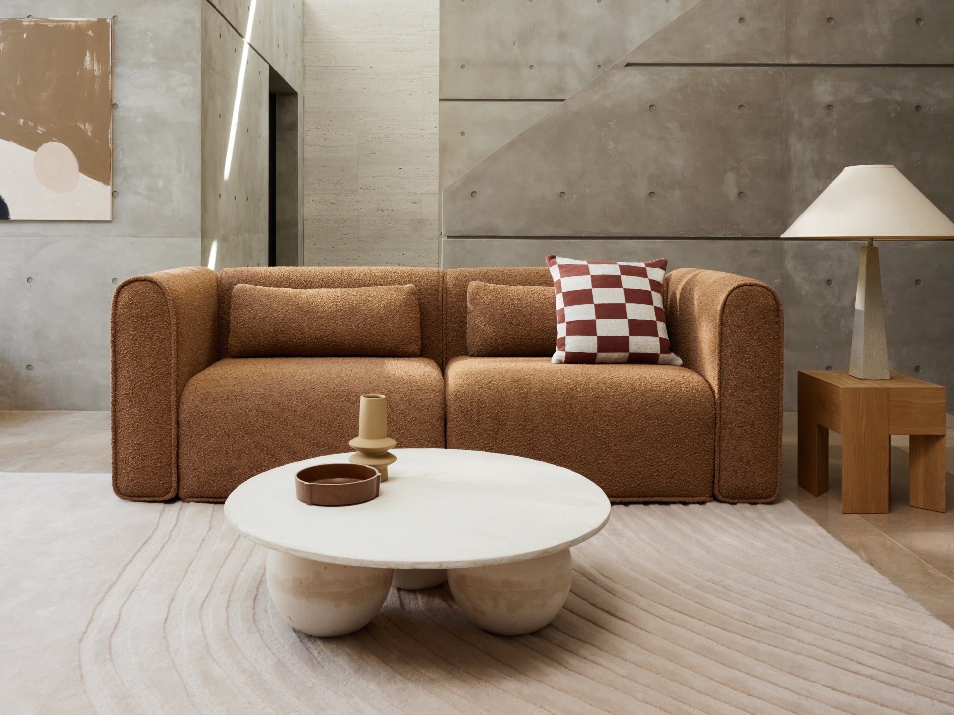 Brown sofa with square arms, two pillows; one solid, one red and white checkered. Round white coffee table and a wooden side table with a lamp.