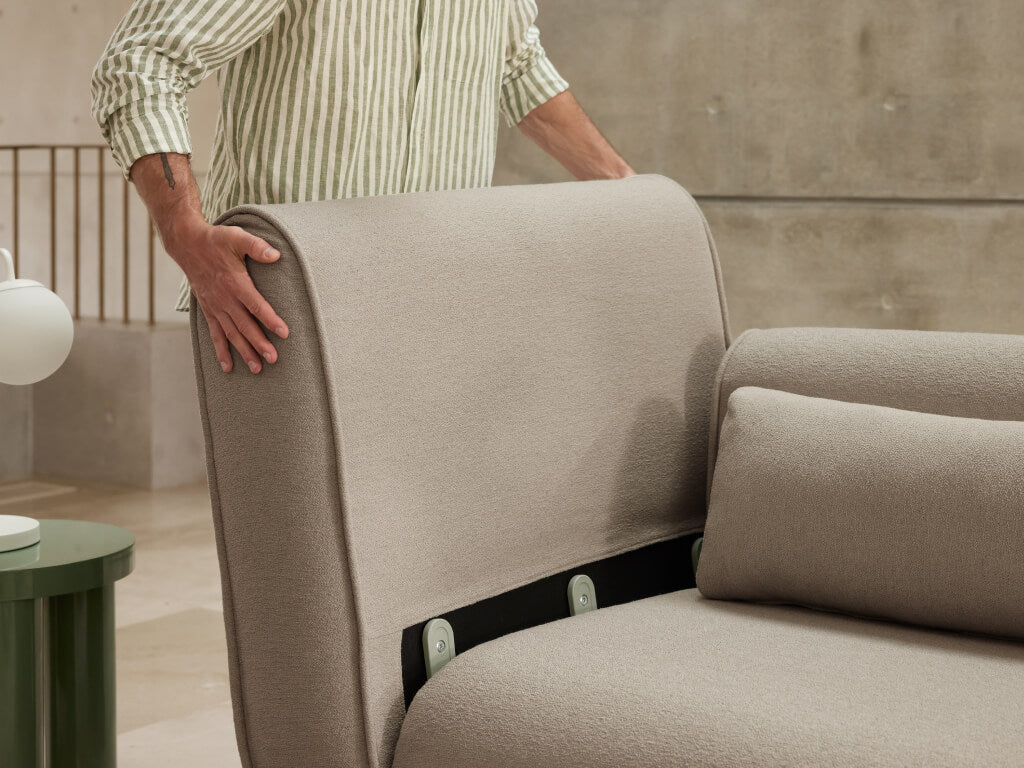 Person adjusting a light brown sofa headrest in a modern minimalist room with green side table and lamp.