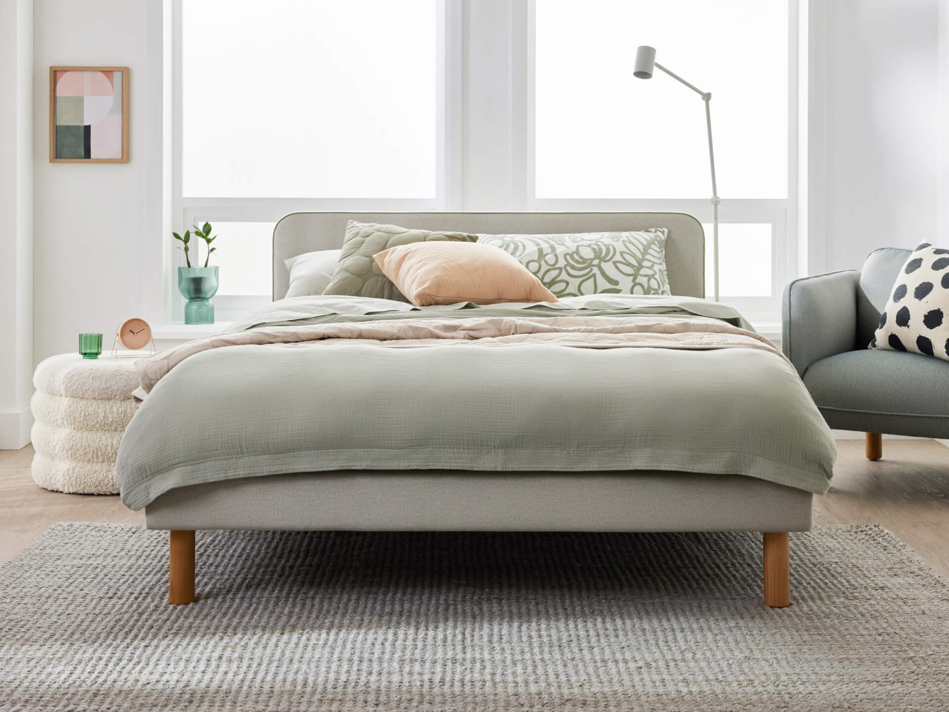 Modern bedroom with a light grey bed, sage green bedding, pastel cushions, a grey rug, white pouffe, and a reading lamp beside a polka dot chair.