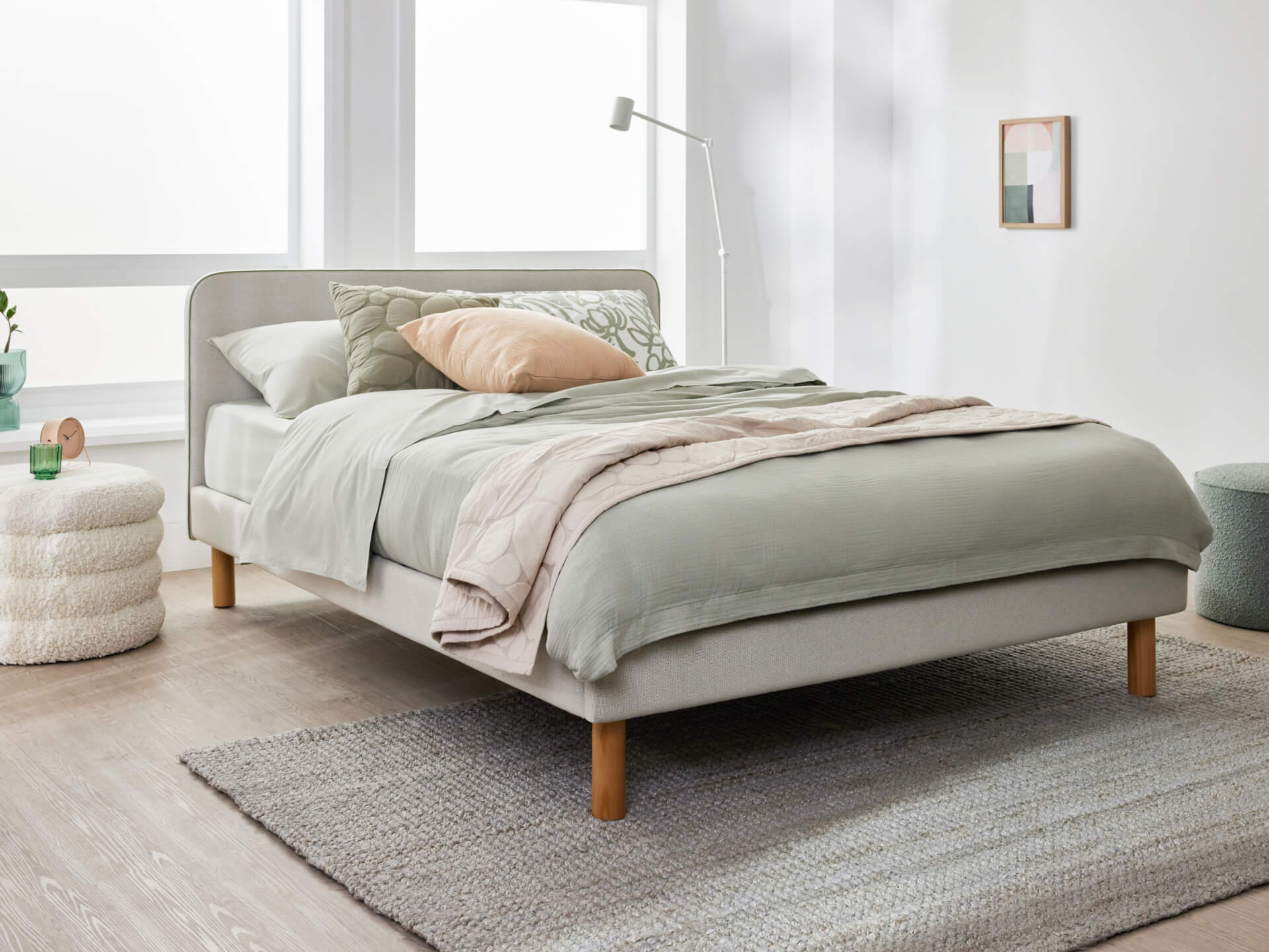 Modern bedroom with a light grey upholstered bed, sage green and peach bedding, on a textured grey rug. Includes a white floor lamp and wall art.