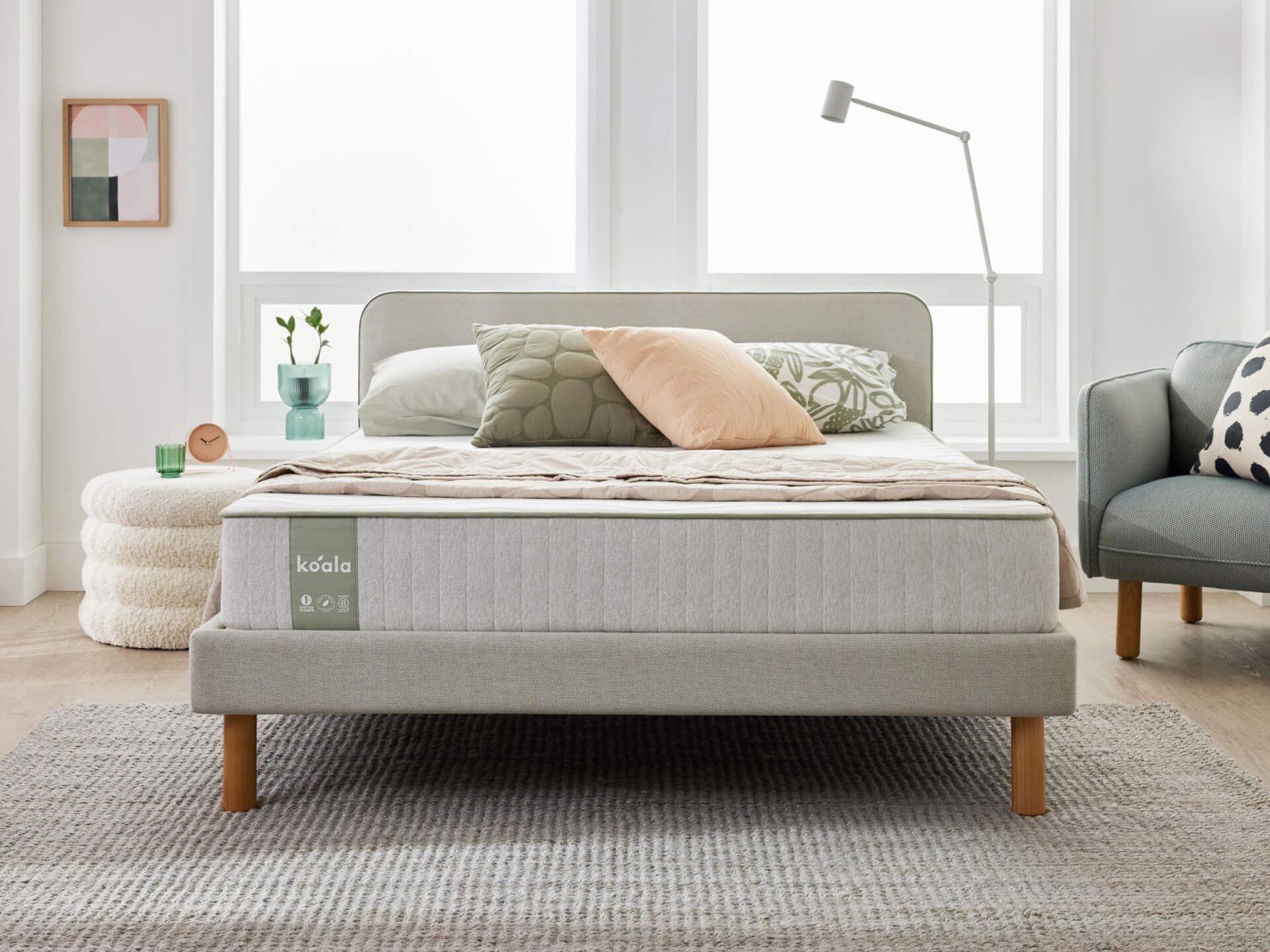 Light grey upholstered bed with wooden legs, topped with pillows and duvet. Nearby, a floor lamp, side table, and a grey chair. Bright room.