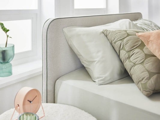 Modern bedroom with a light grey upholstered bed, green and peach cushions, and a round bedside clock. Green vase on the windowsill.