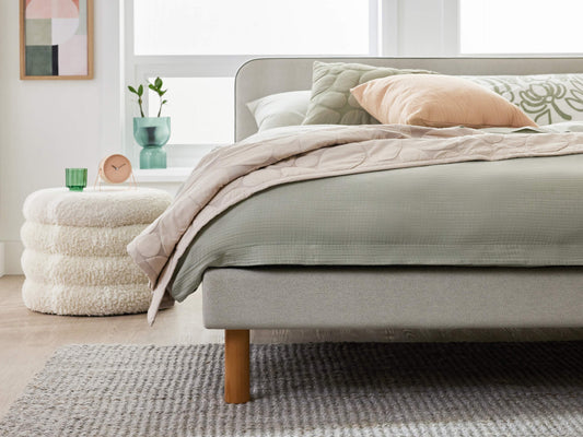 Modern bedroom with grey bed, wooden legs, and green bedding. Textured grey rug, cream ottoman, and pastel cushions enhance the decor.