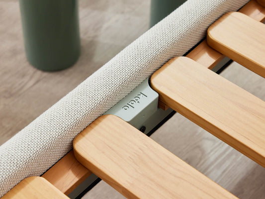 Close-up of a wooden bed slat frame with light wood slats and a beige fabric edge; label reads "Kodable" on a metal part.