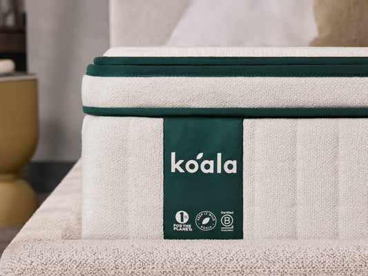 Close-up of a cream mattress with green edging and a "koala" label, resting on a textured base. Background features a blurred beige chair and a round table.