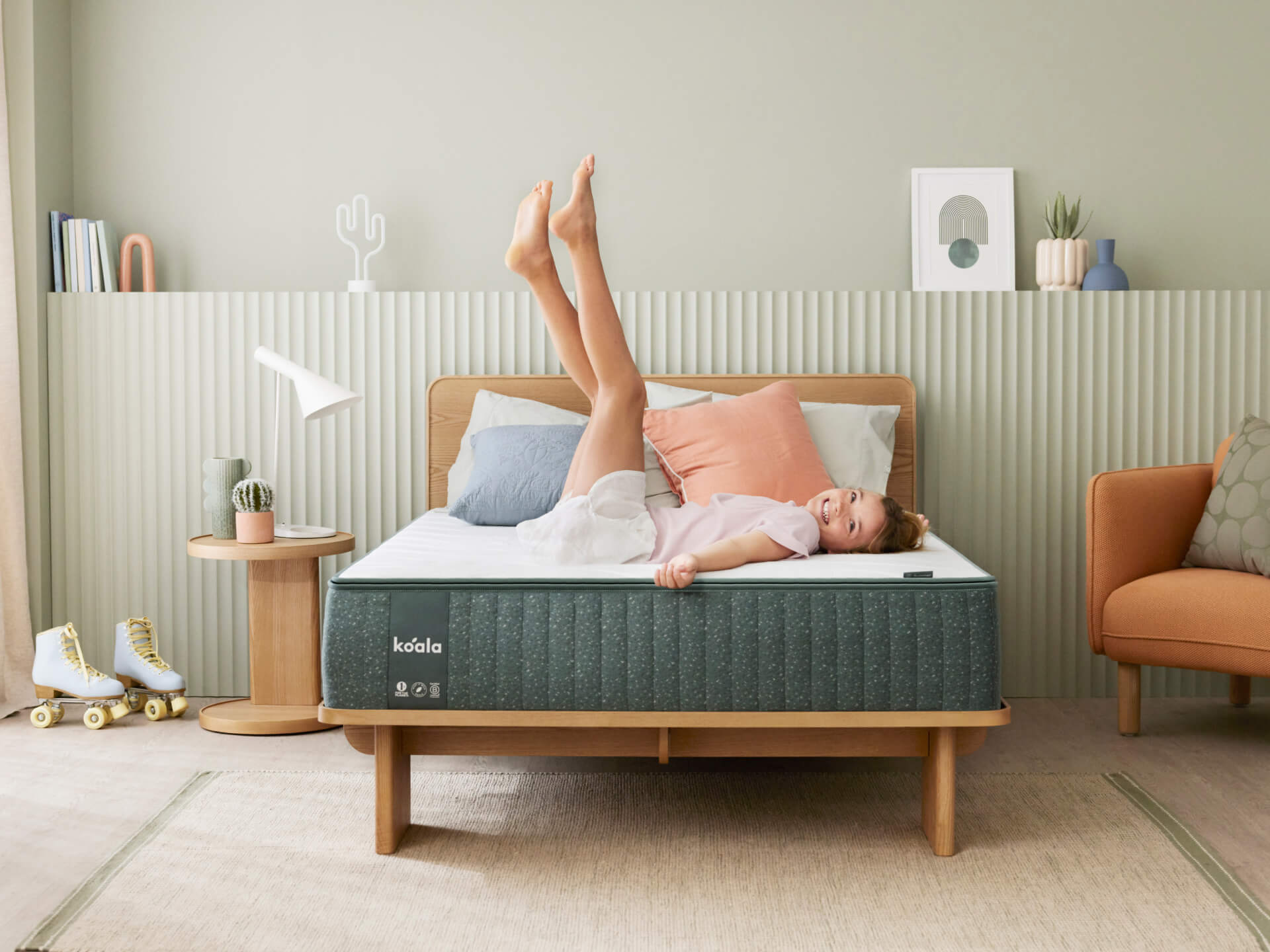 Child on teal mattress, wooden bed frame, pastel cushions, and a bedside table with a lamp. Green wall, decor items, and roller skates nearby.