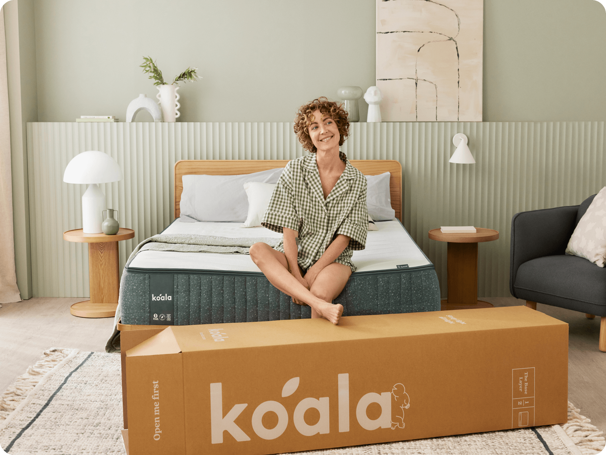 Smiling woman sitting cross-legged on a bed with a Koala mattress and unpacked Koala mattress box in front.