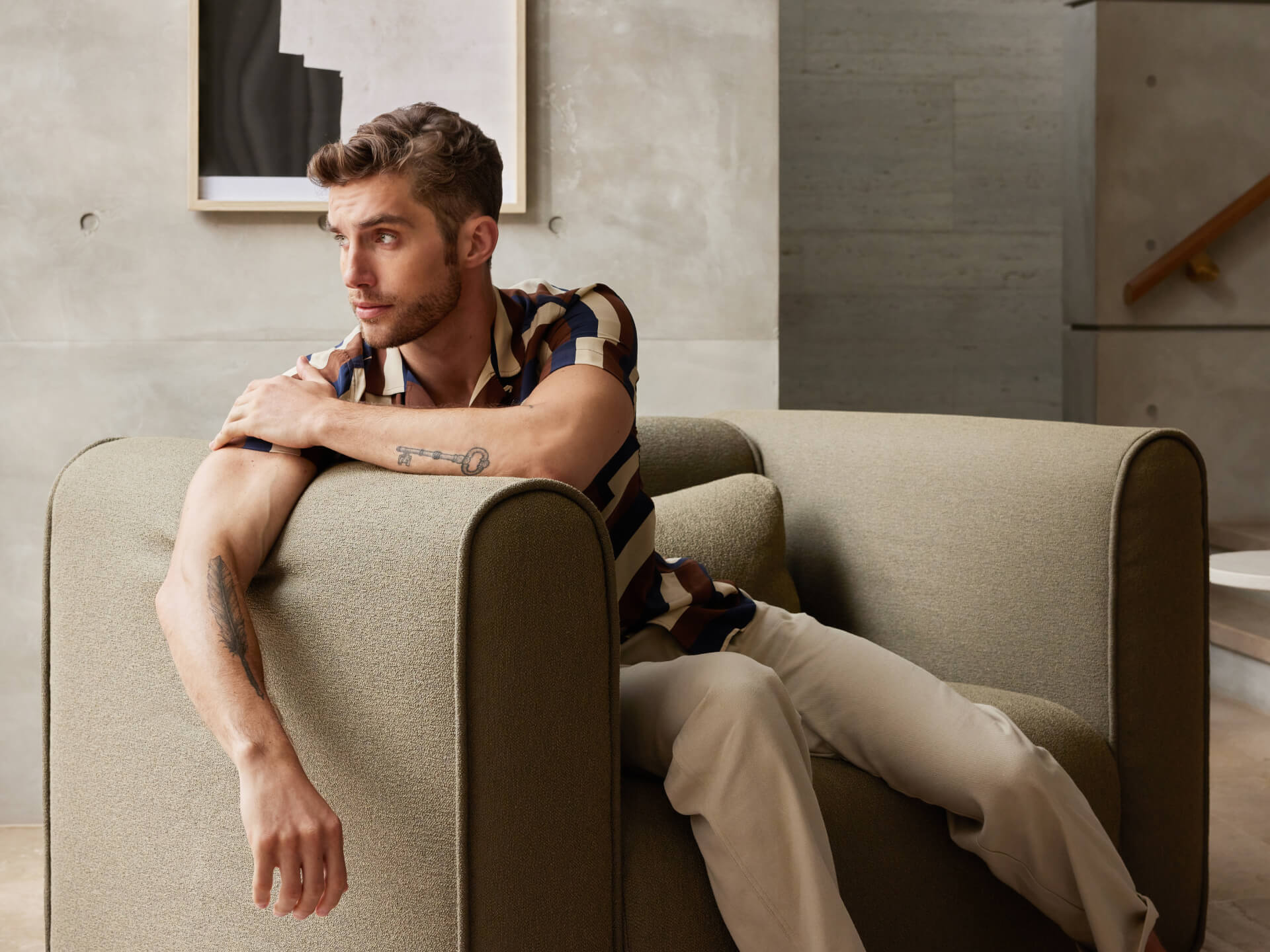 Man sitting on a light olive armchair in a modern living room, wearing a patterned shirt and white trousers, with minimalistic decor.