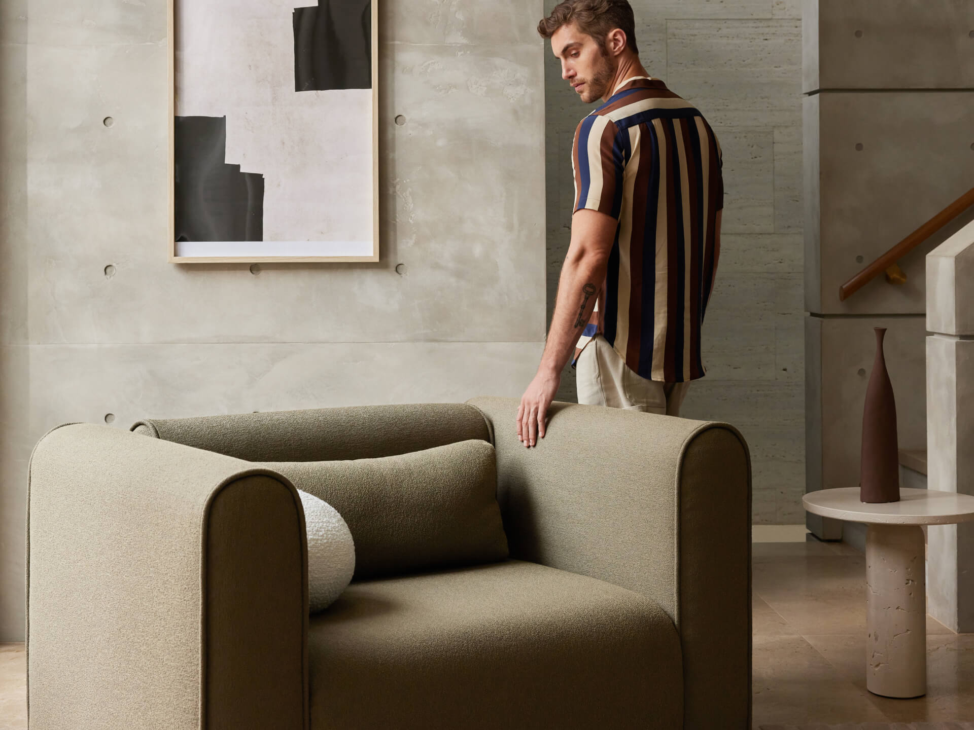 Olive green armchair with round cushions in a modern living room. A man in a striped shirt stands nearby. Grey wall with abstract art and a round table.