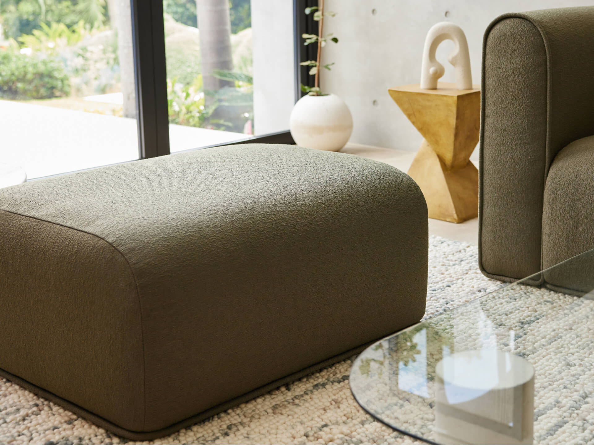 Olive green ottoman on a textured light-coloured rug, next to a rounded armchair and glass coffee table. Modern decor with natural light.