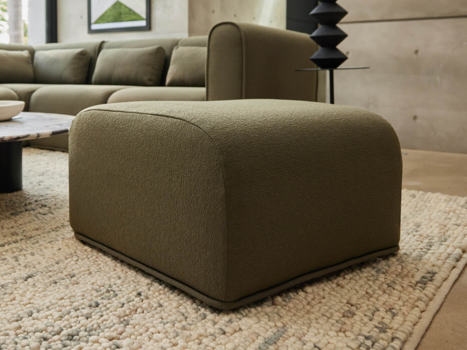 Olive green ottoman on a beige textured rug, near a modern dark grey lamp and a sectional sofa. Concrete walls in the background.