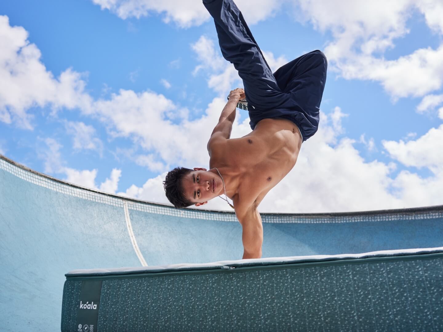 Man performing a handstand on a green mattress outdoors, under a blue sky with clouds.