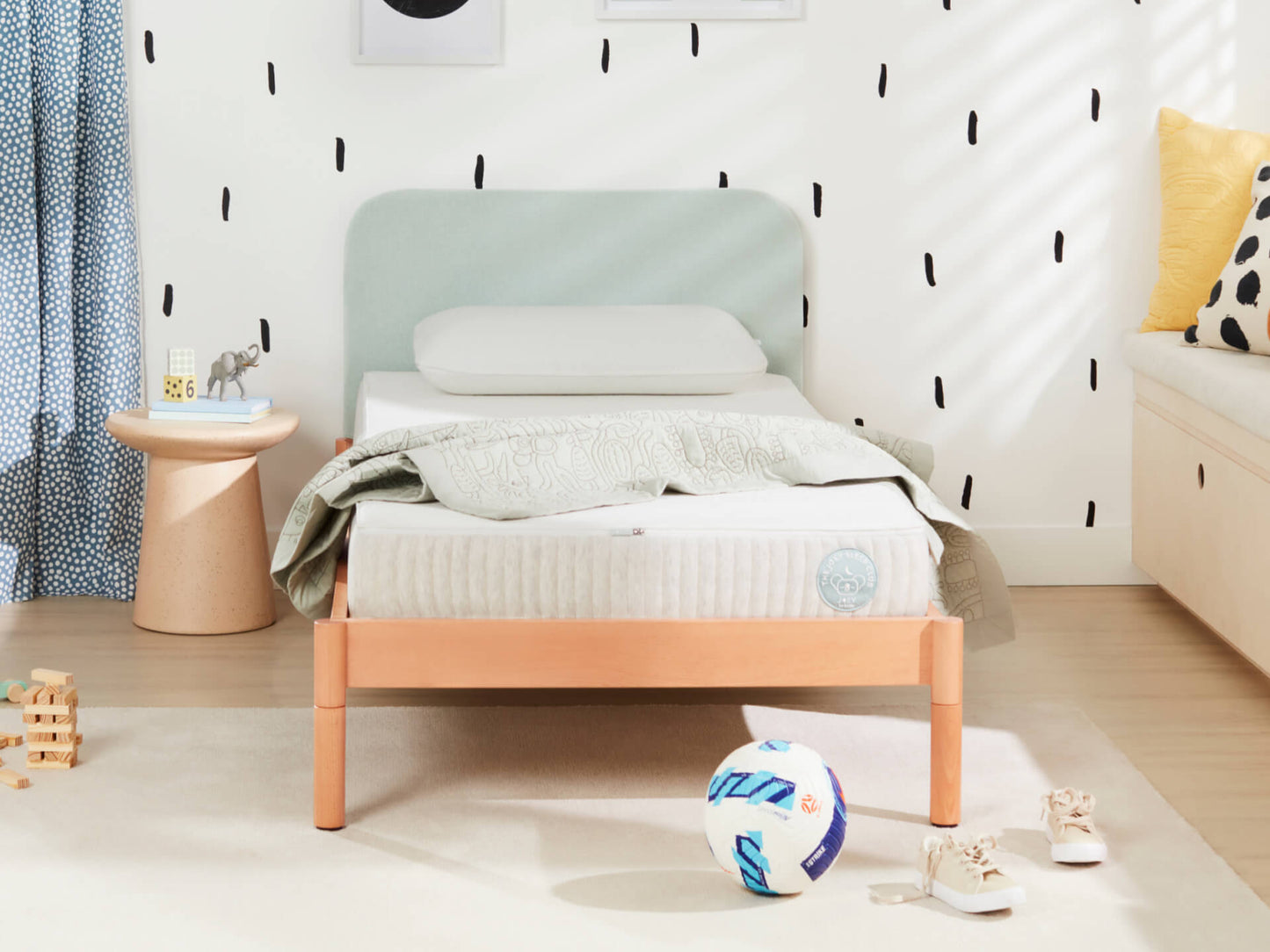 Child's bedroom with a wooden single bed, light blue headboard, polka dot wallpaper, small round table, ball, and sneakers on light wood floor.