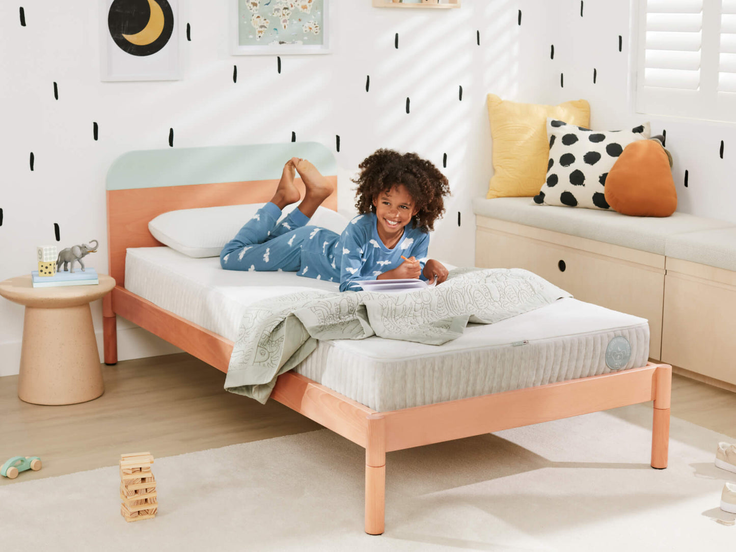 Child on light wood bed with white mattress, pastel headboard, in a bright room. Cushioned bench with pillows; small side table with toy.