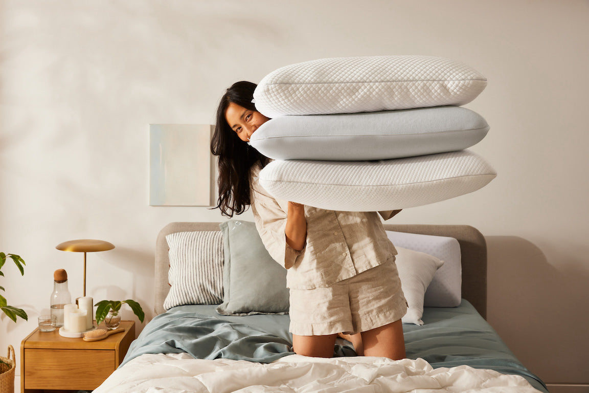 Woman in beige pyjamas holding three pillows on a bed. Bedside table with lamp and plants in the background.