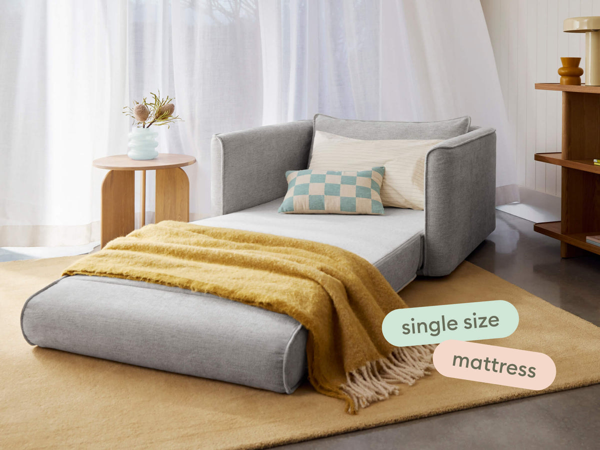 Single-size grey sofa bed with a plush mattress, smooth fabric upholstery, and rounded edges. Set in a modern living room with neutral decor and soft lighting.