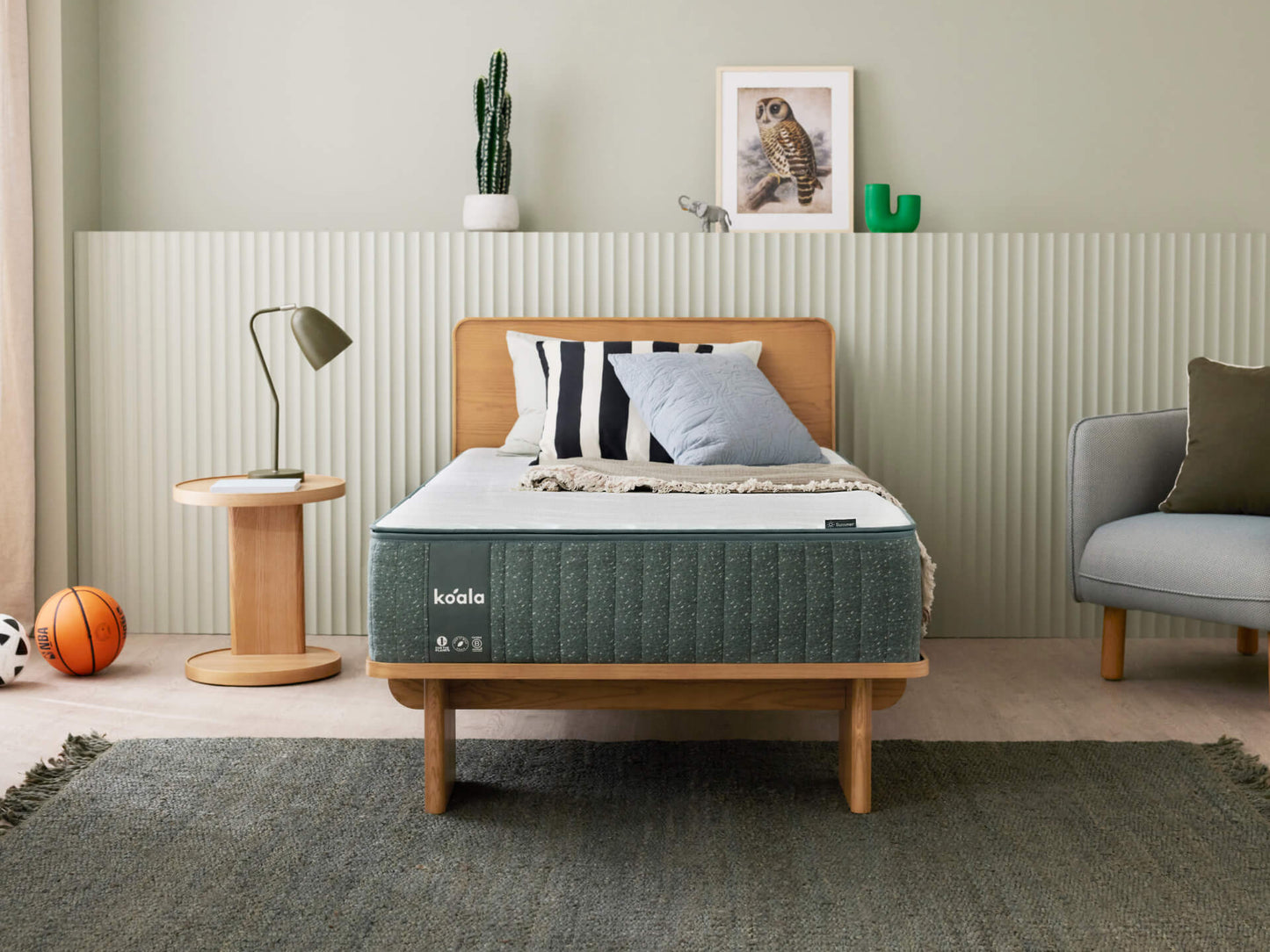 Bedroom with a wooden bed frame, green mattress, and striped pillows. A side table with a lamp, grey armchair, and decor complete the scene.