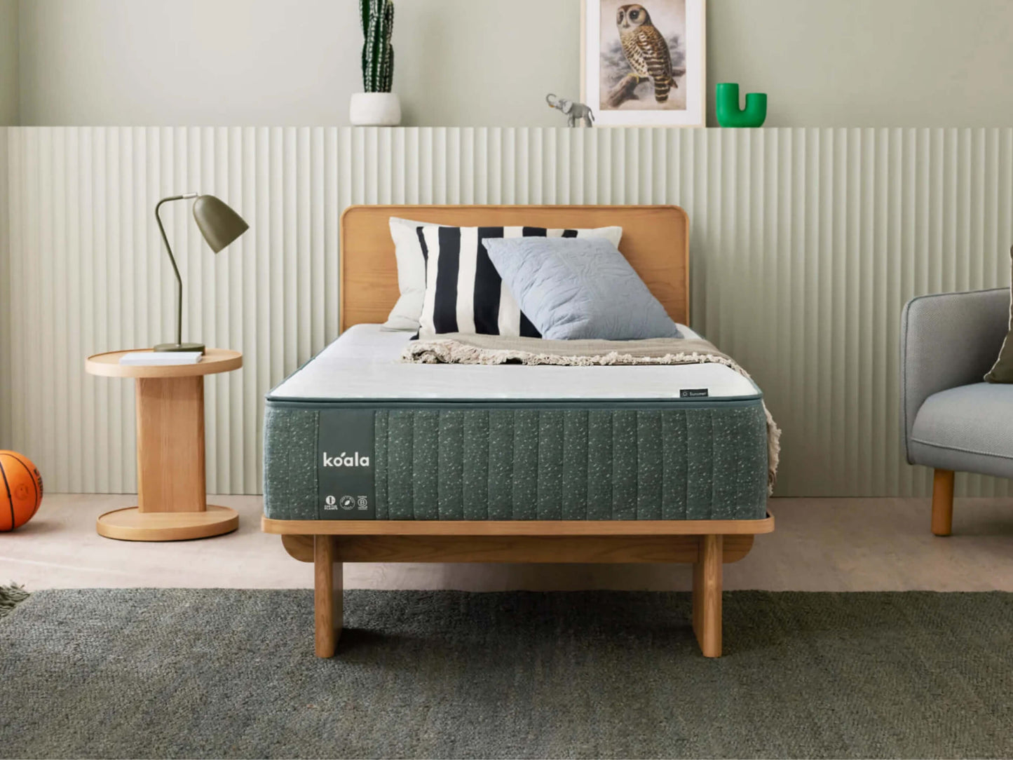 Modern bedroom with a wooden bed featuring a grey mattress, light blue pillows, and a striped cushion. Nearby are a wooden side table, lamp, and grey chair.