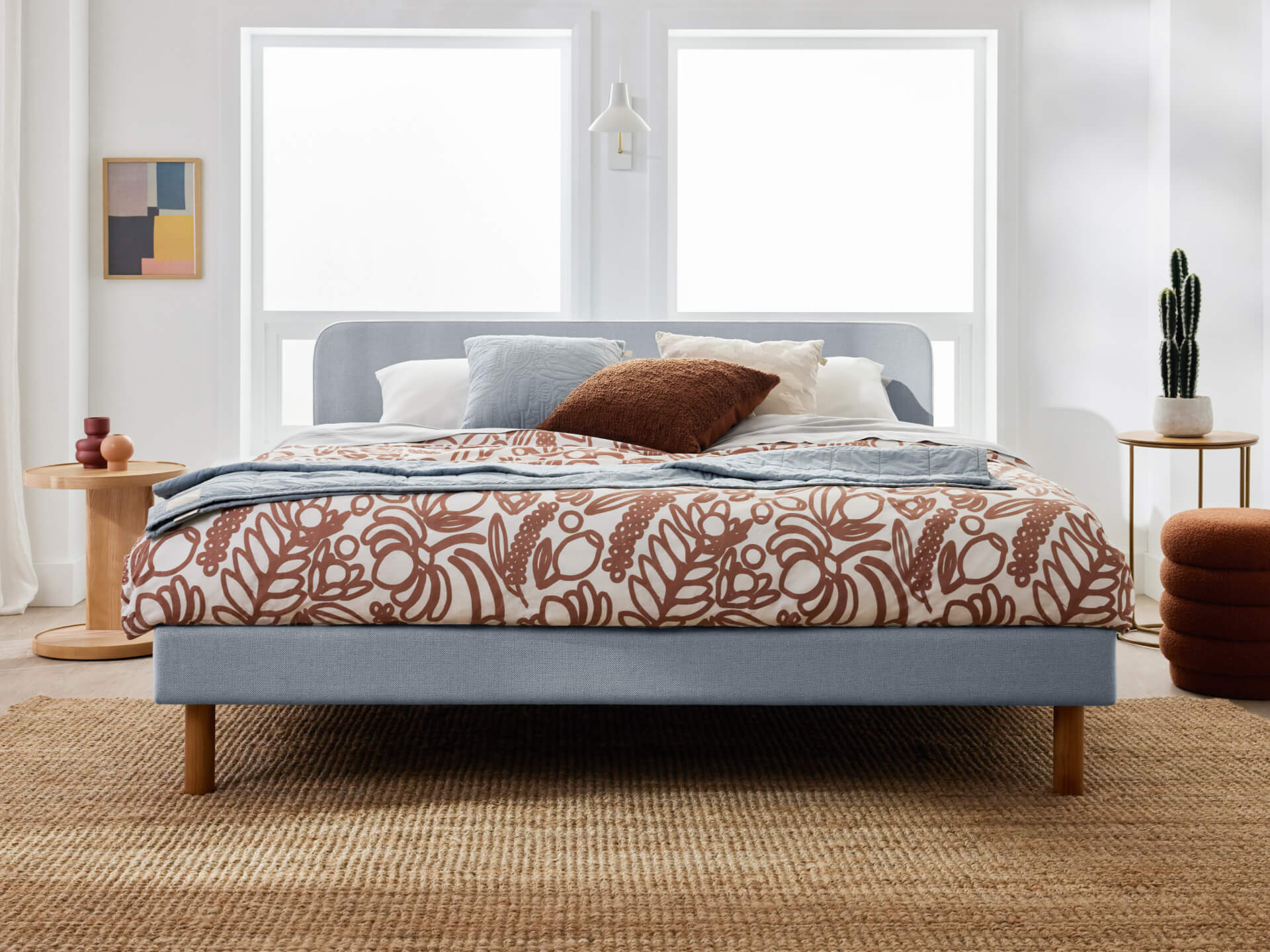 Light grey bed with patterned brown and white duvet, blue and brown cushions. Natural wood bedside tables, cactus plant, jute rug. Bright room.