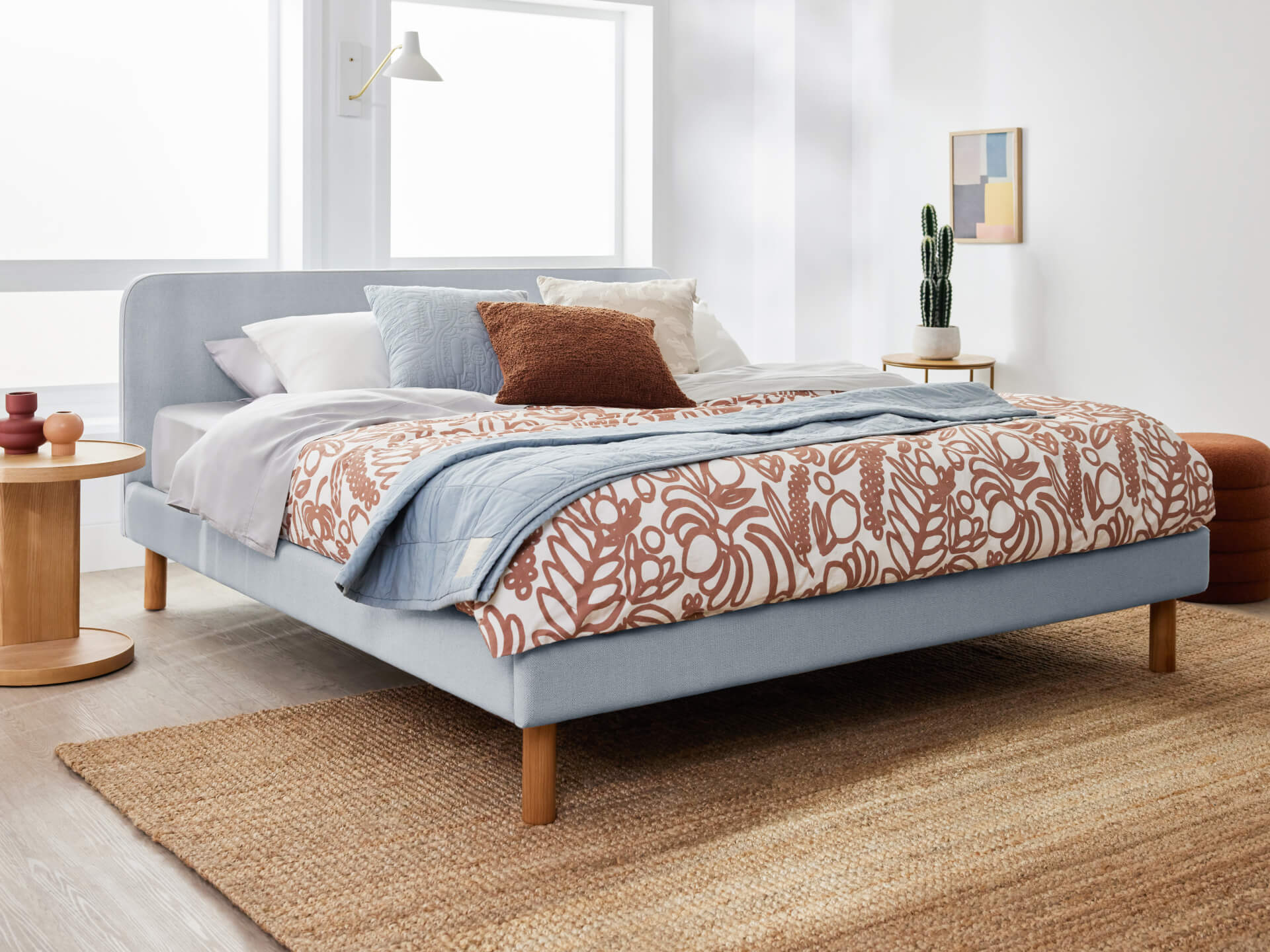 Modern bedroom with a light blue upholstered bed, floral patterned quilt, wooden side tables, and a jute rug. Scandi decor with a cactus and artwork.