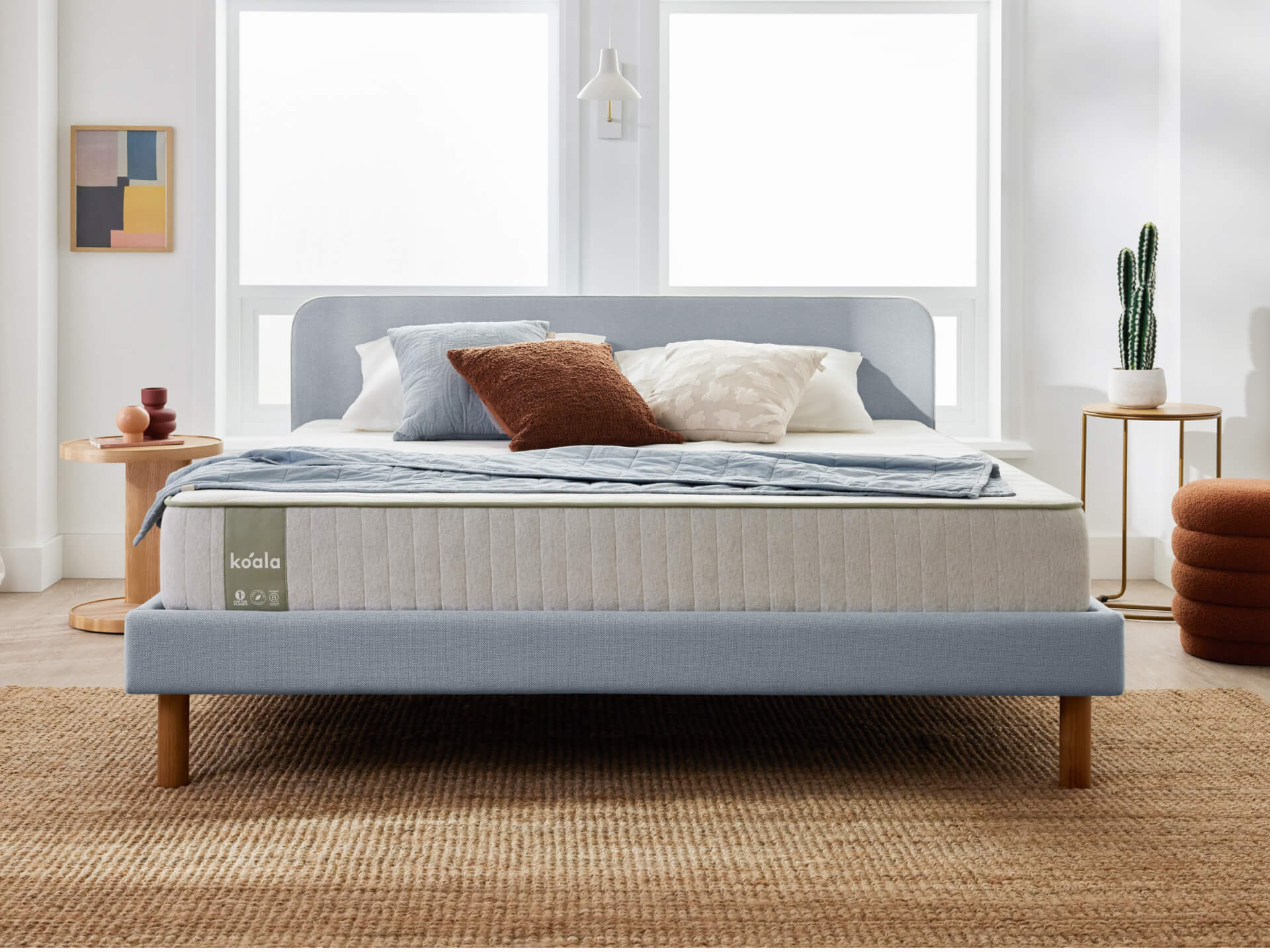 Bedroom scene with a light blue bed frame, white mattress, various cushions, a wooden side table, a rug, and decorative items in a bright room.