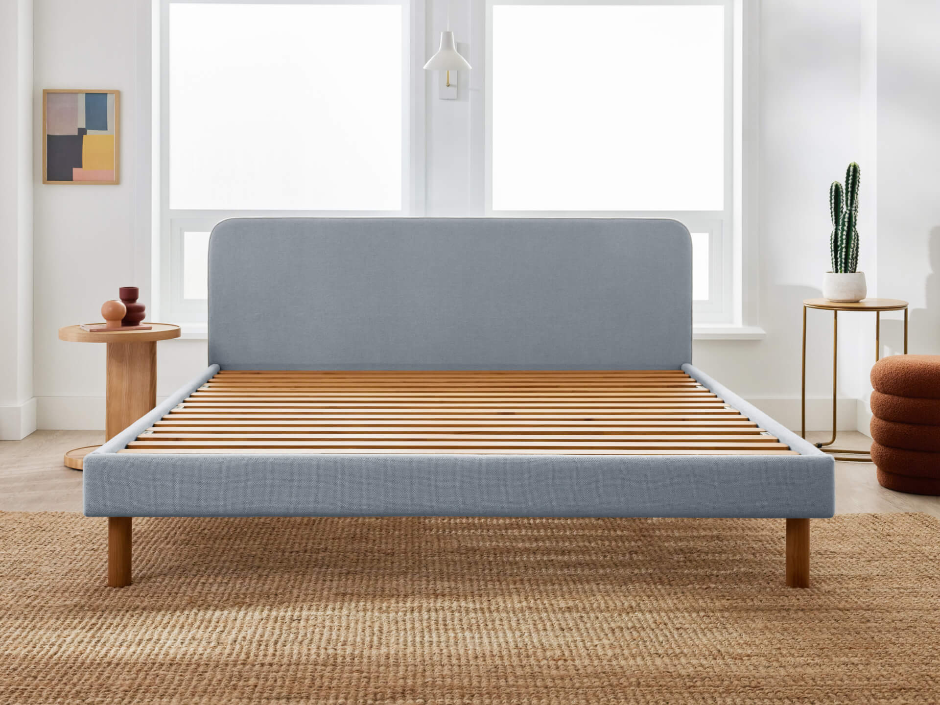 Light grey bed frame with wooden slats in a modern bedroom, featuring a rug, small round tables, decor, and a cactus.