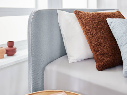 Light grey upholstered bed with a white pillow, brown textured cushion, and light blue pillow. Wooden table and pottery visible beside window.