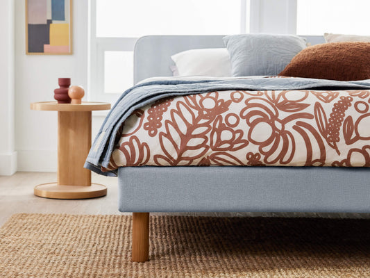 Modern bedroom with light grey upholstered bed, brown patterned duvet, and neutral pillows. Wooden side table and textured rug on light floor.