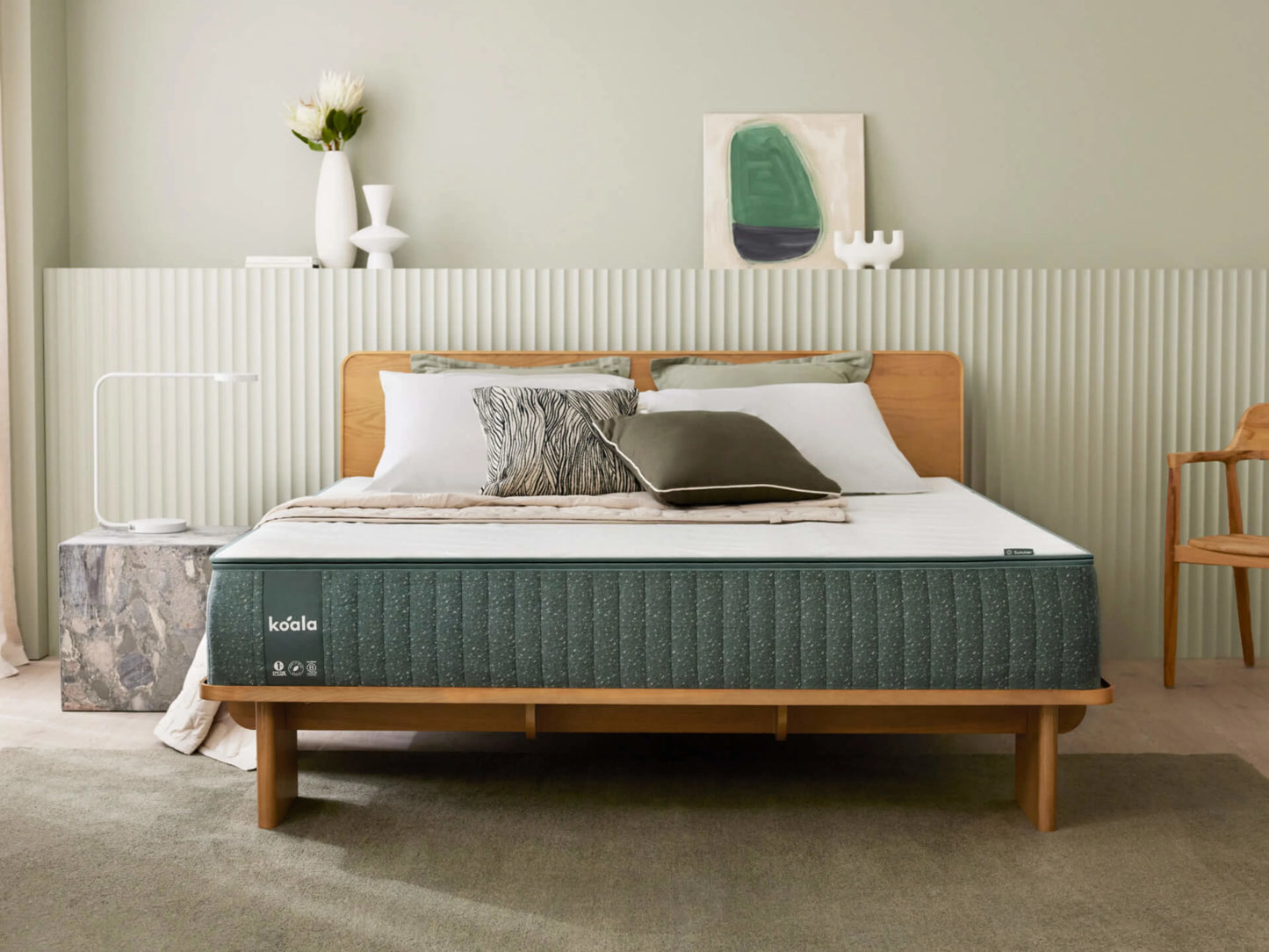 Modern bedroom with a thick mattress on a wooden bed frame. Light grey walls, a stone side table, white lamp, and green decor accents.