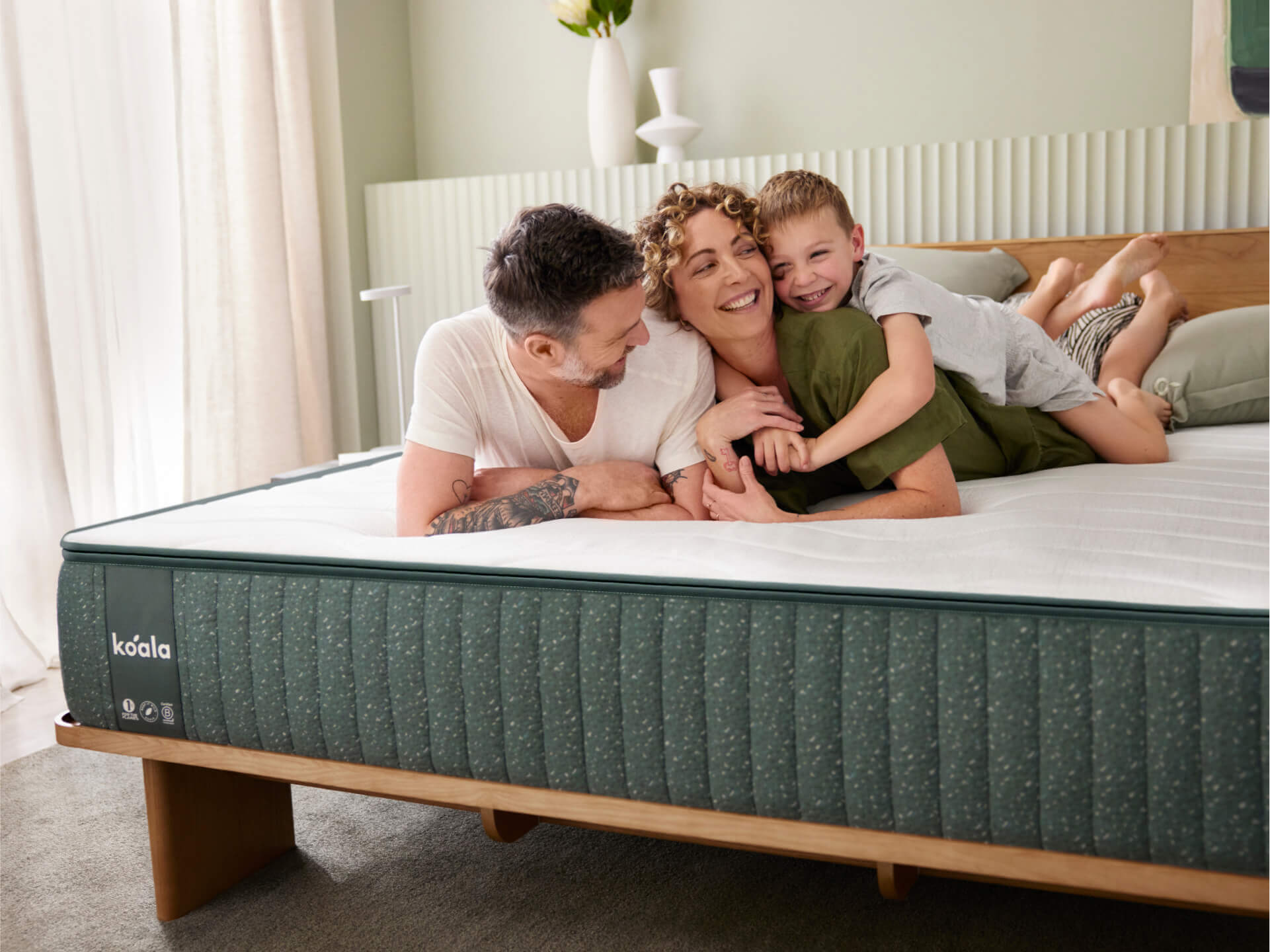 Family smiling on a bed with a dark green mattress in a bright room. Upholstered headboard and soft carpet add warmth to the decor.