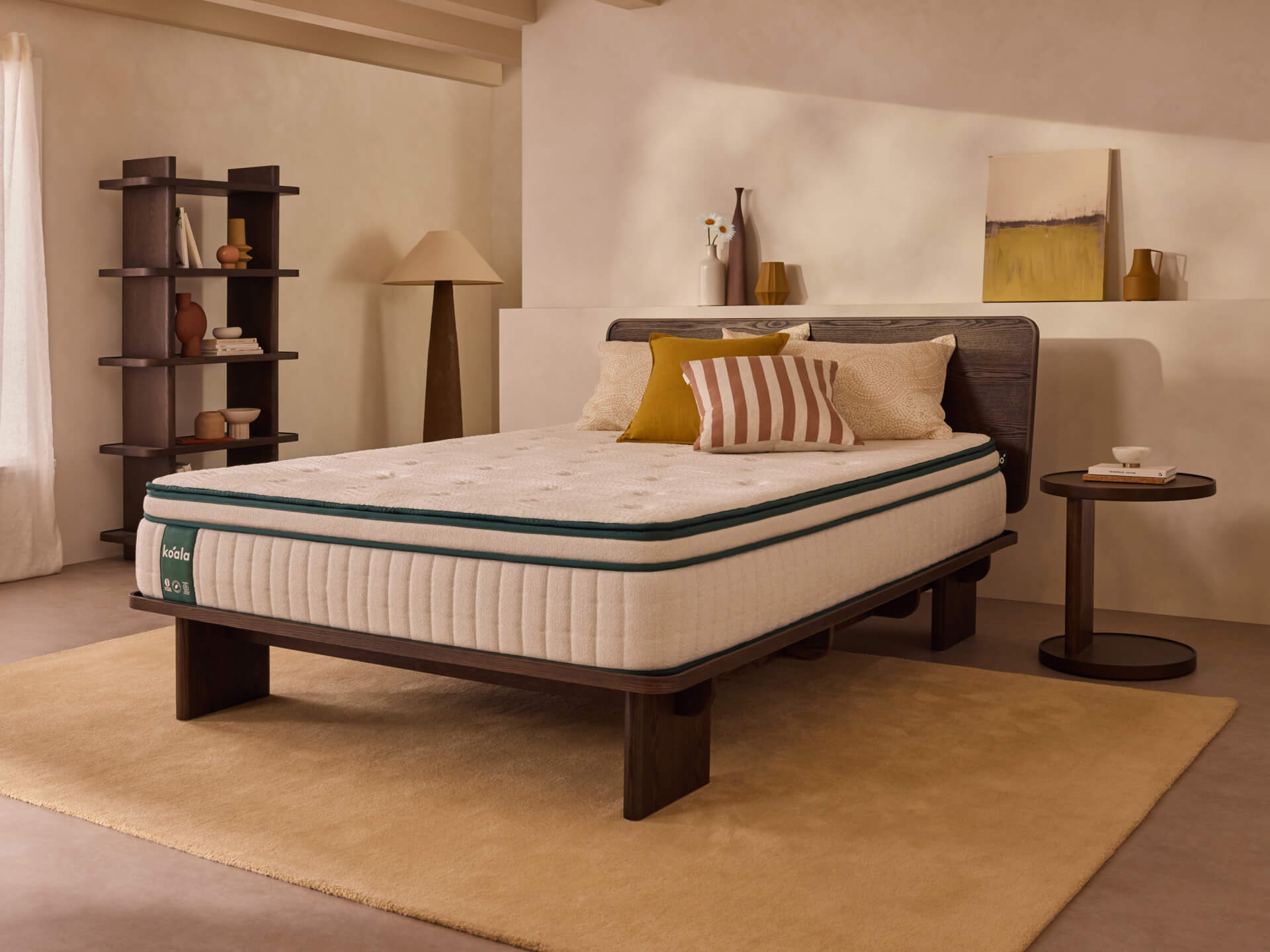 Modern bedroom with a queen-sized mattress on a wooden bed frame, beige and mustard cushions, side table, and minimalist decor.
