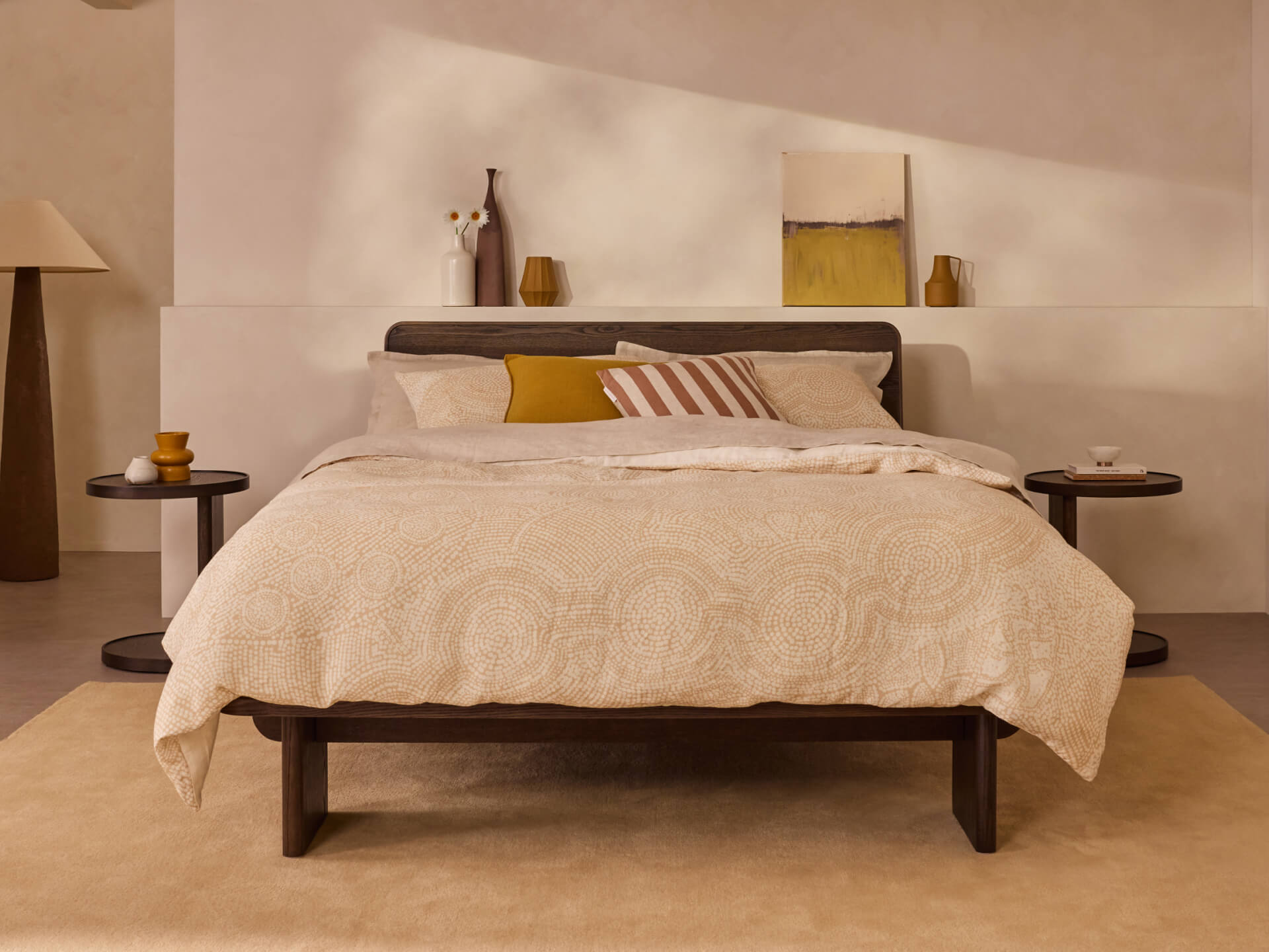 Bedroom with a dark wood bed frame, beige patterned bedding, a mustard cushion, and flanked by round side tables. Decor includes a lamp and art.