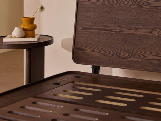 Dark brown wooden bed frame with slatted base next to a round side table holding a yellow vase, white cup, and books.