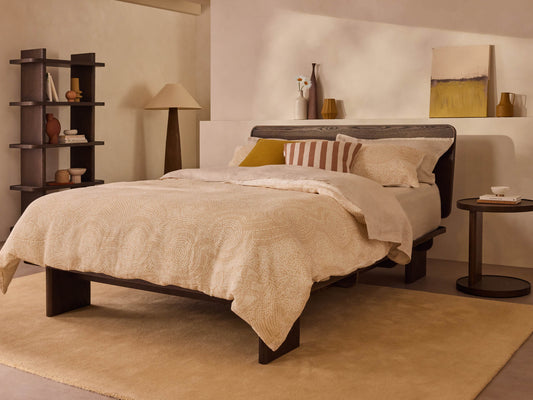 Stylish bedroom with a double bed featuring cream bedding, wooden frame, round side table, tall lamp, and modern bookshelf against beige walls.