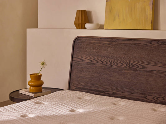 Wooden bedhead with grain pattern, cream textured bedspread, brown side table with mustard vase, white bowl, and a book. Minimalist decor.