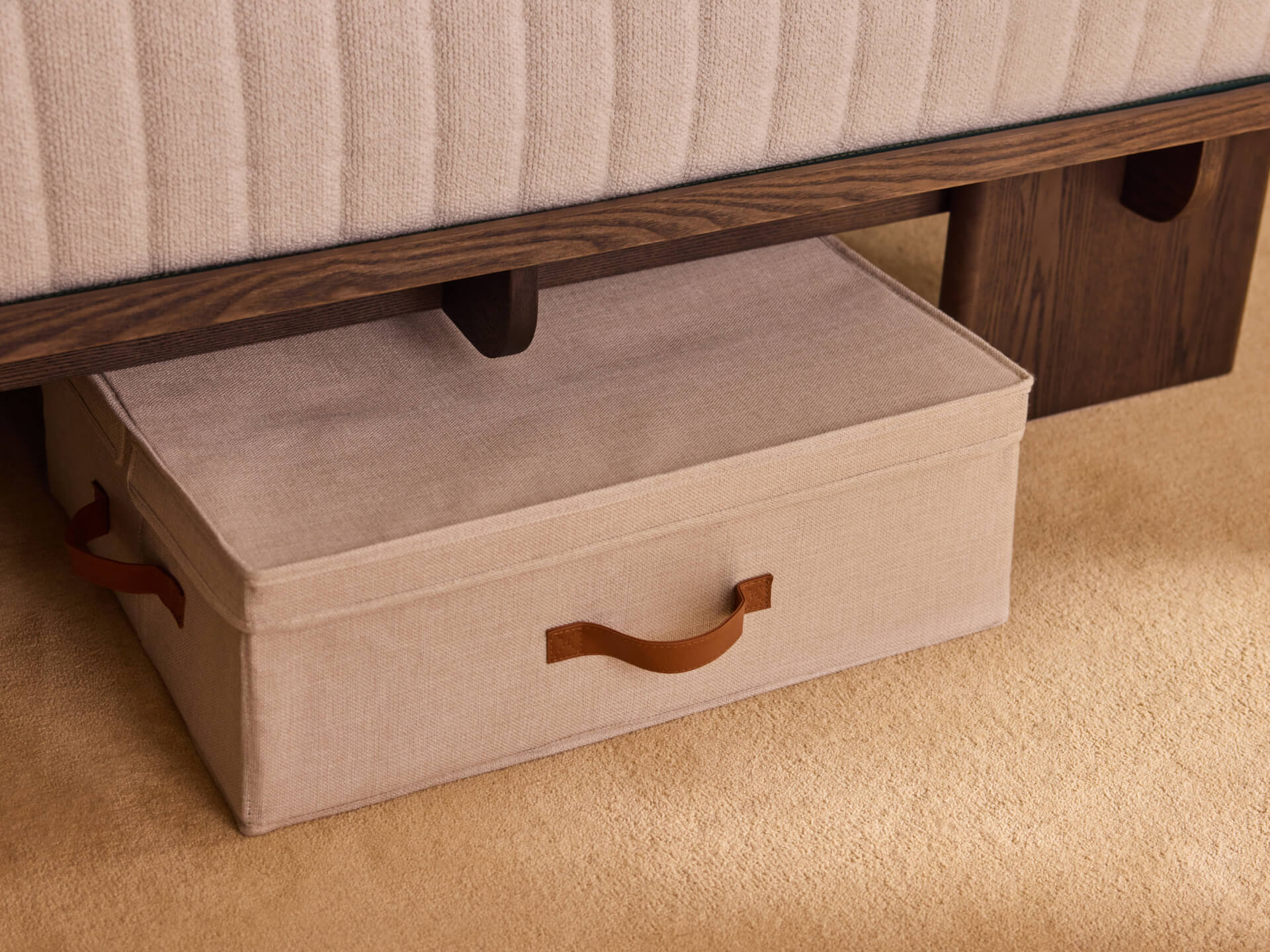 Fabric storage box with brown leather handles under a bed with a light brown mattress, on a beige carpet.