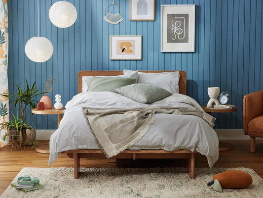 Bedroom with a wooden bed, light grey bedding, green cushions, blue panelled wall, assorted wall art, and indoor plants.