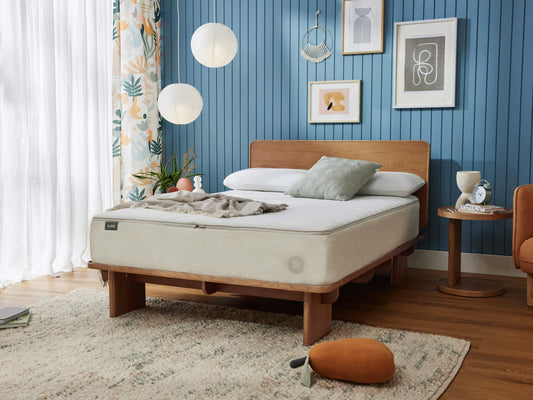 Modern bedroom with wooden bed frame and mattress, blue panelled wall, patterned curtains, round wooden bedside table, and abstract wall art.