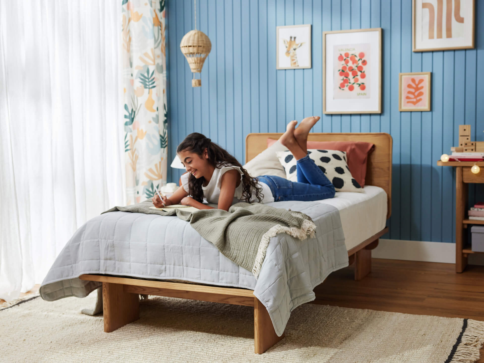 A girl lies on a wooden single bed with grey and green blankets in a cosy, blue-panelled room. Framed art and patterned curtains adorn the walls.