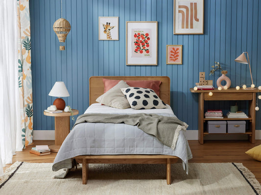 Cosy bedroom with a wooden bed, light grey quilt, spotted pillow, wooden bedside table, blue panelled wall, and framed artwork.