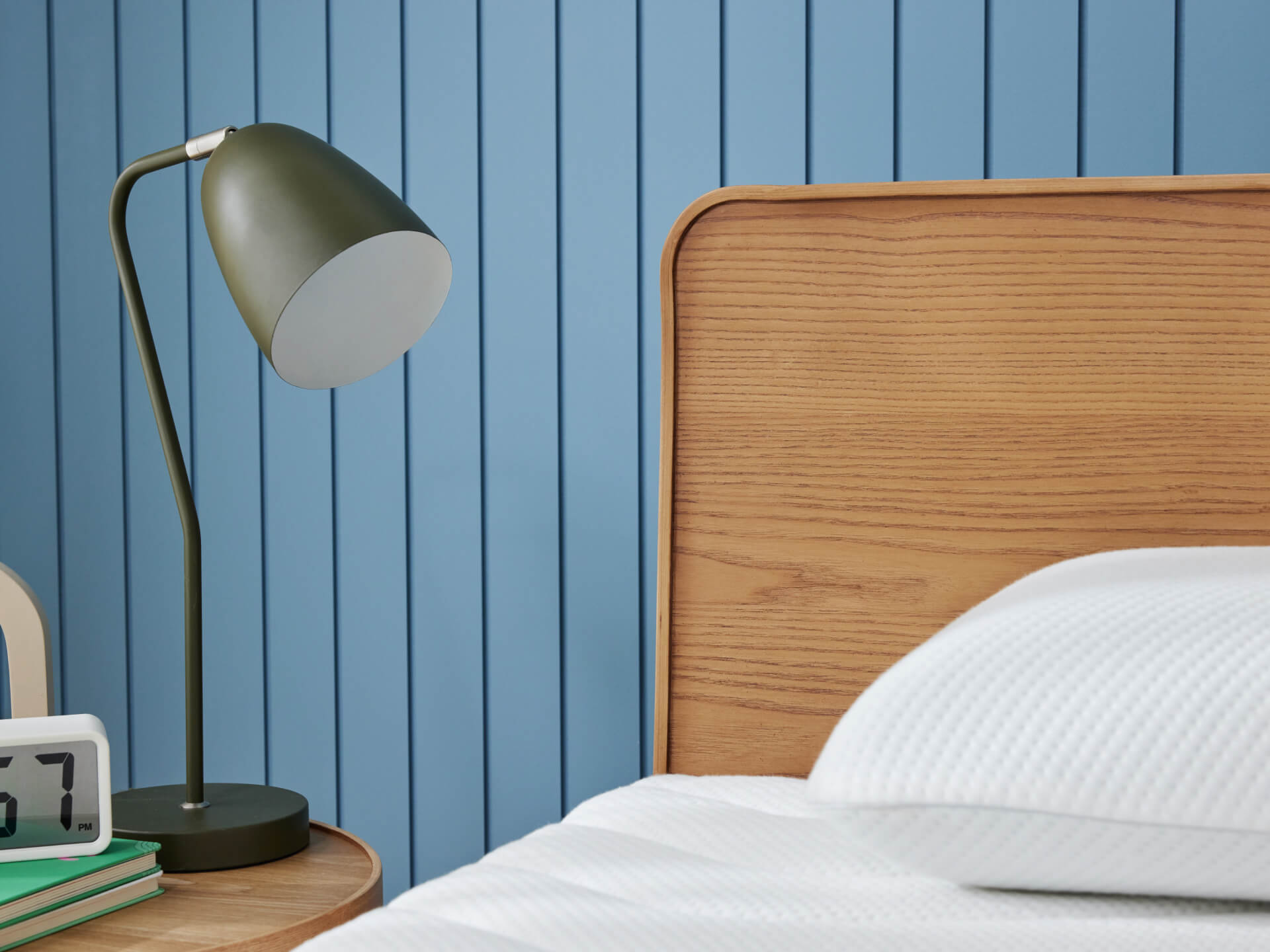 Bed with wooden headboard, white pillow, green desk lamp on a round table, clock, and books against a blue panelled wall.
